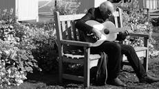一位老人在户外木凳上弹吉他，这张照片是黑白的