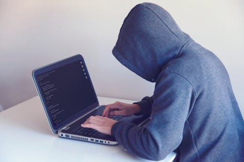 Free Side view of unrecognizable hacker in hoodie sitting at white table and working remotely on netbook in light room near wall Stock Photo