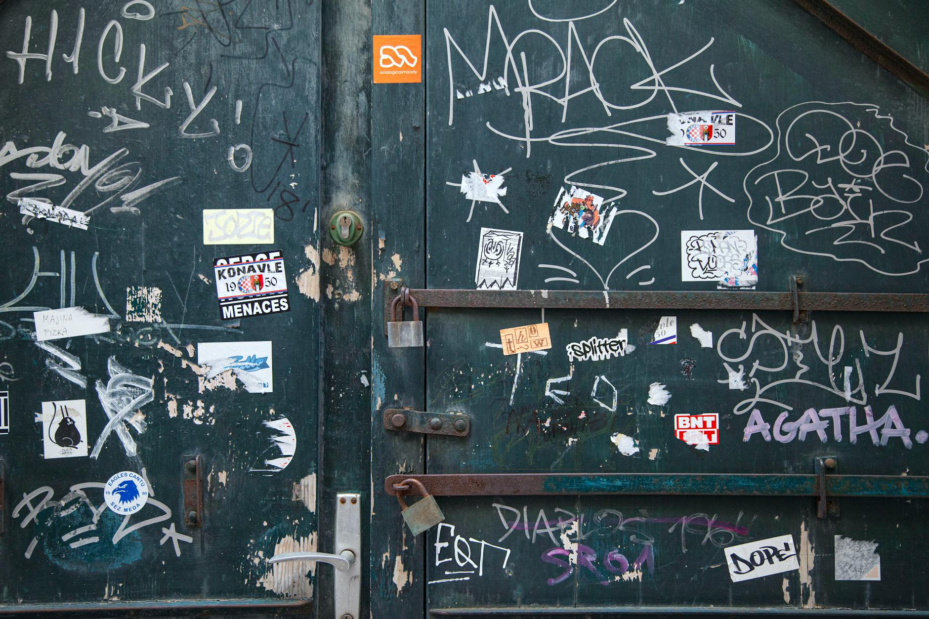 Weathered metal door with locks and doorhandle near white painted graffiti and small stickers in daylight