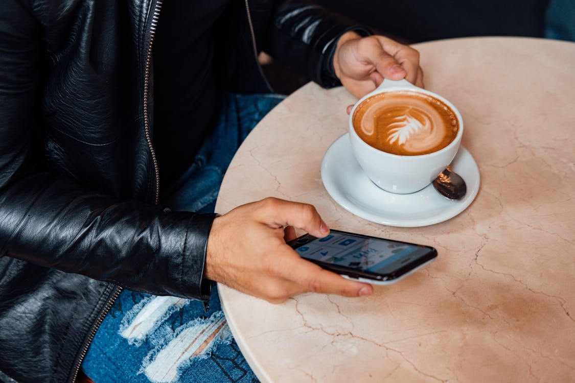 Free Person Holding White Ceramic Mug With Brown Liquid and a Smartphone Stock Photo