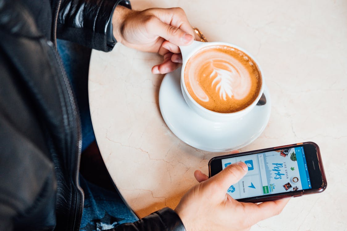 Free Person Holding White Ceramic Cup While Using a Smartphone Stock Photo
