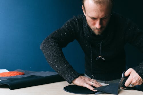 Concentrated male tailor with scissors cutting round shaped piece of leather while working with fabric in atelier