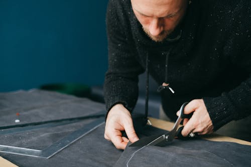 Serious male tailor with scissors cutting circle out of leather material while standing at workbench in atelier