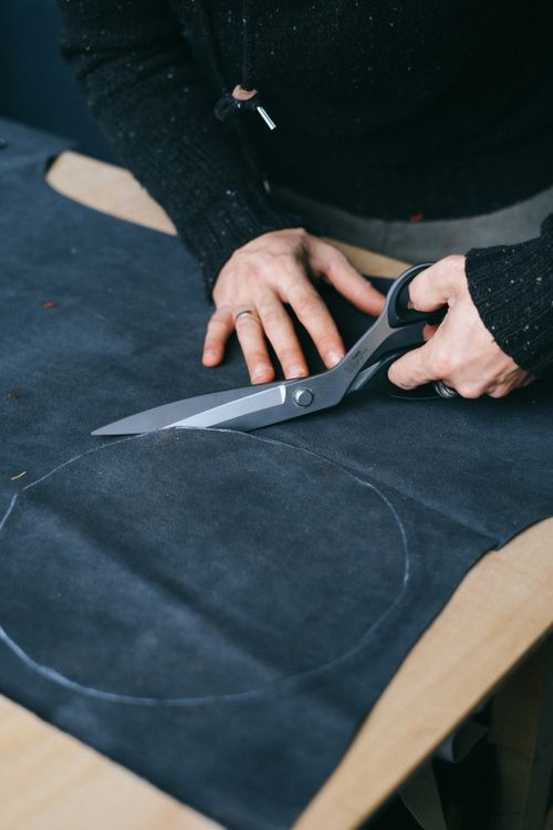 Tailor cutting dark fabric with large shears at a workshop cutting table, representing the fabric cutting stage of plush production