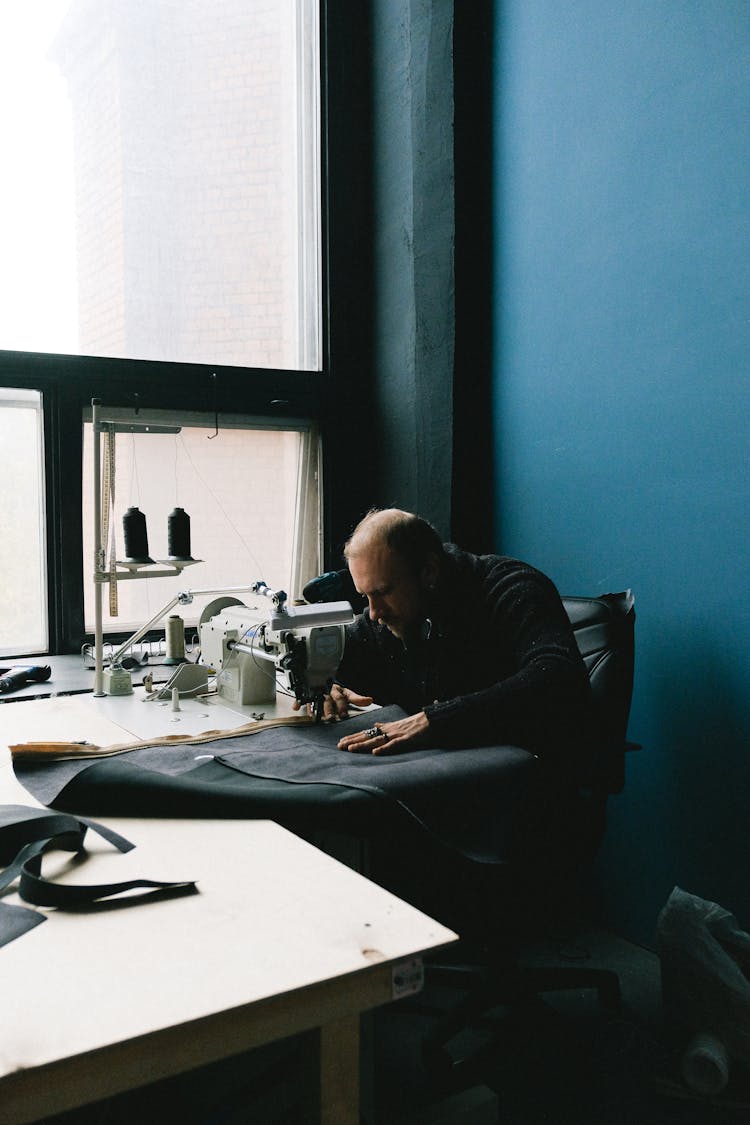 A Man Sitting And Sewing A Fabric On A Wooden Table