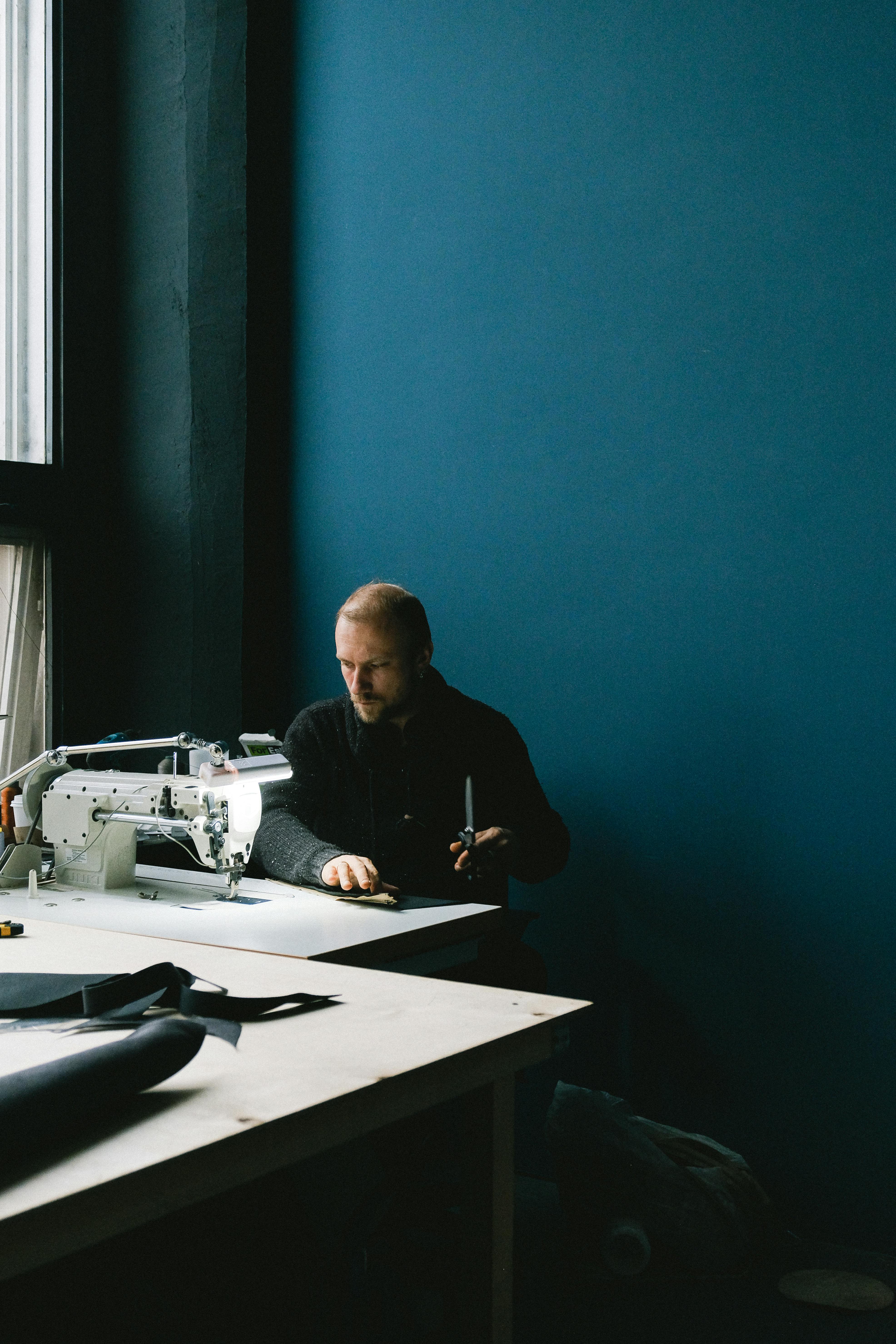 Free Focused tailor working on a sewing project in a dimly lit workshop with a blue wall backdrop. Stock Photo