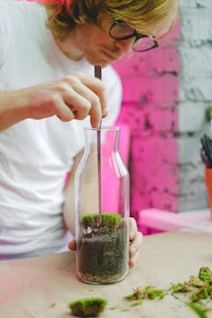 A man carefully builds a terrarium in a vase using soil and moss under pink lighting.