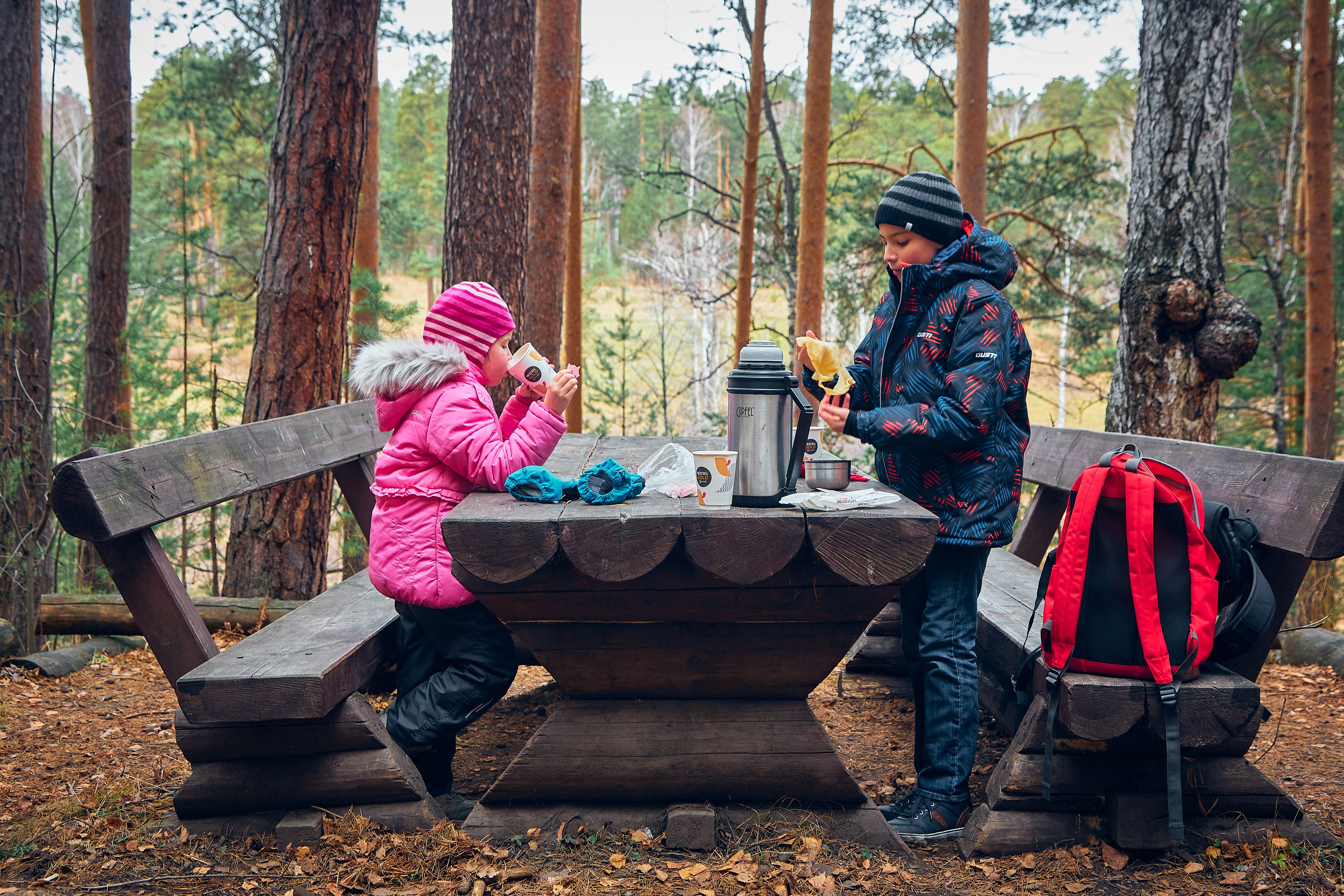 Two children enjoy a picnic in a forest, dressed in winter clothing, highlighting outdoor fun.