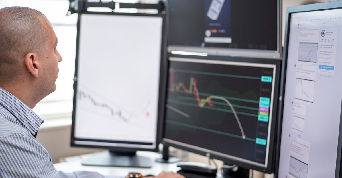 Stock trader analyzing financial graphs on multiple computer monitors in an office setting.