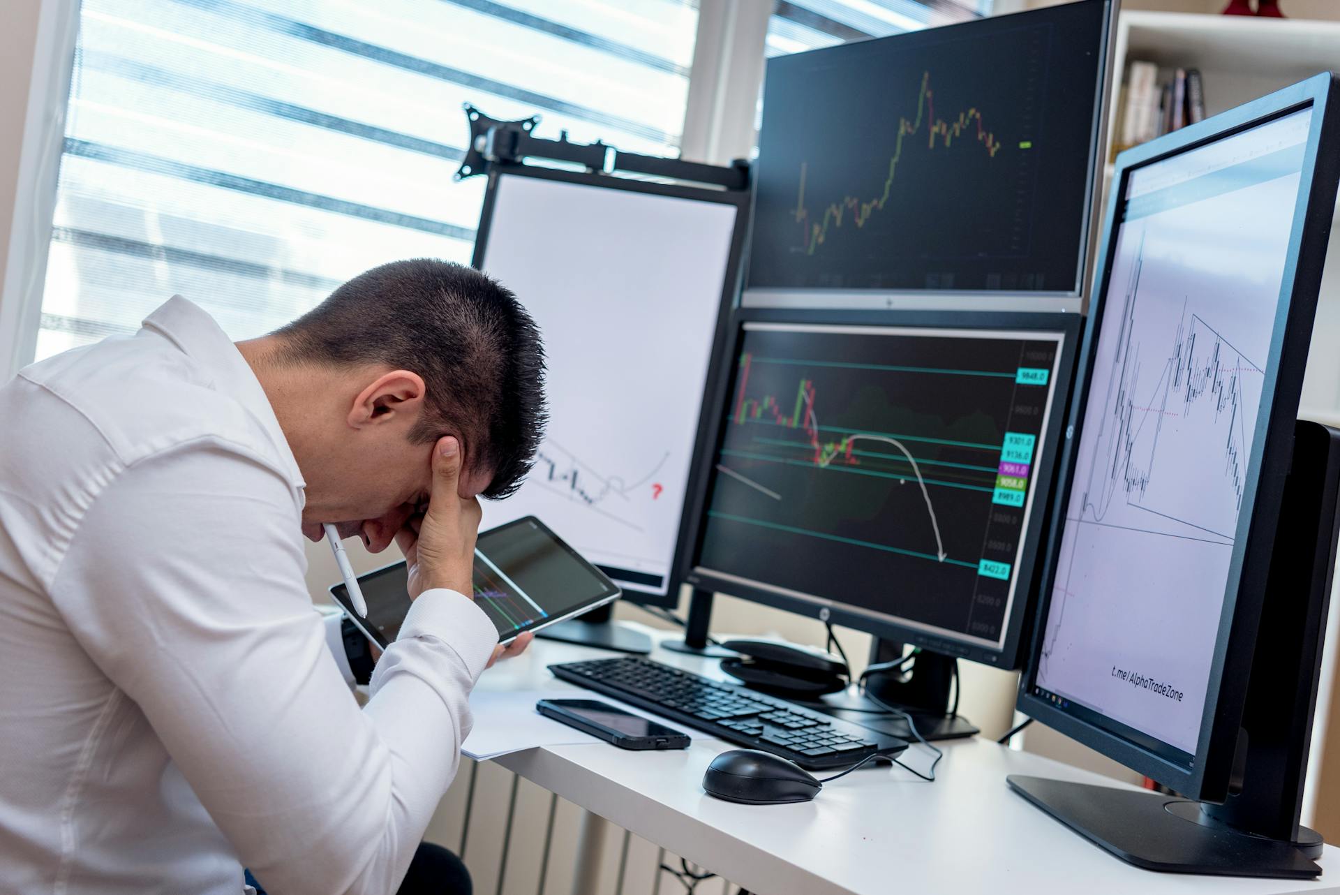 A stressed trader in an office setting analyzes market data on multiple monitors using a tablet.