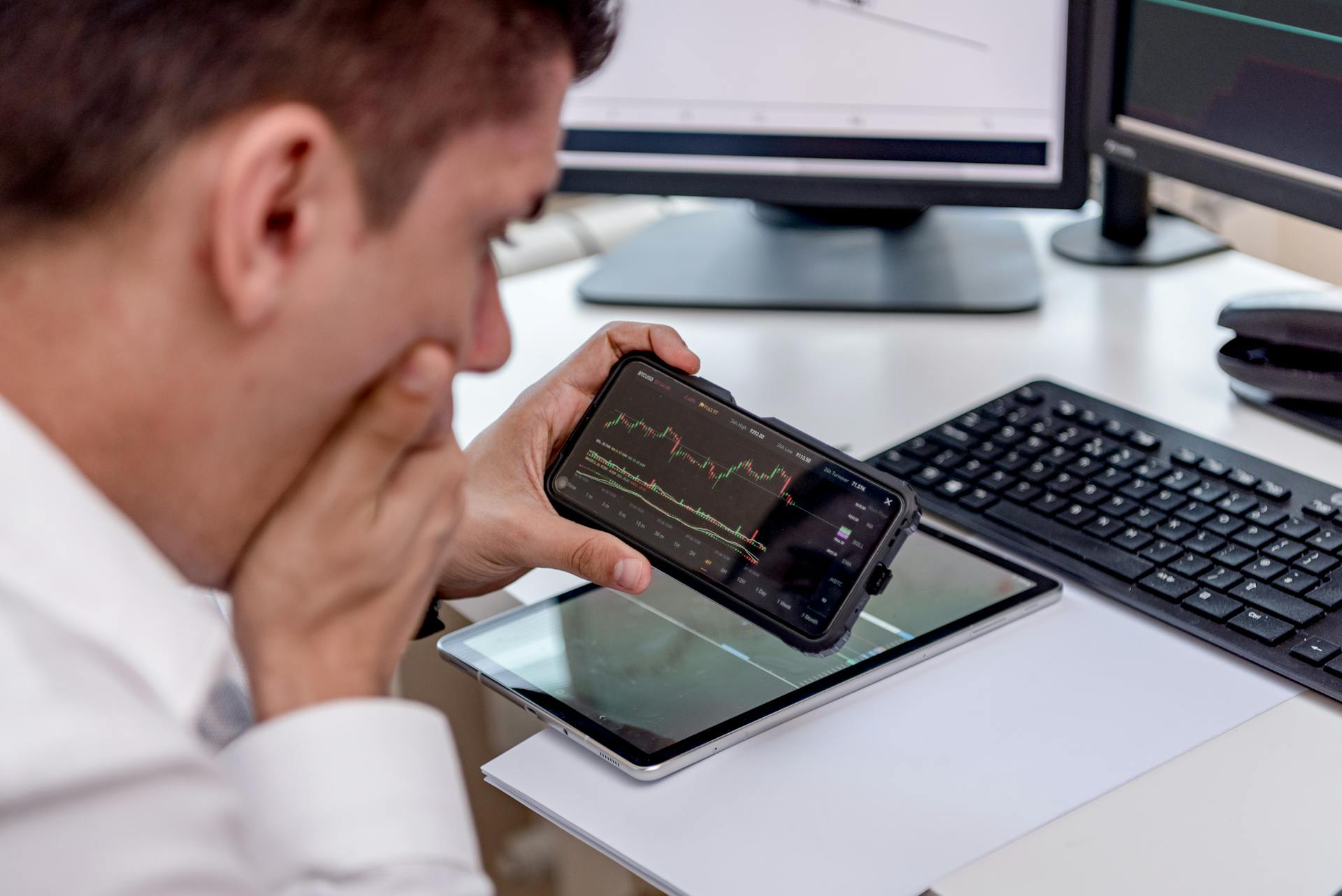 Businessman focuses on stock market analysis using smartphone and digital devices in modern workspace.