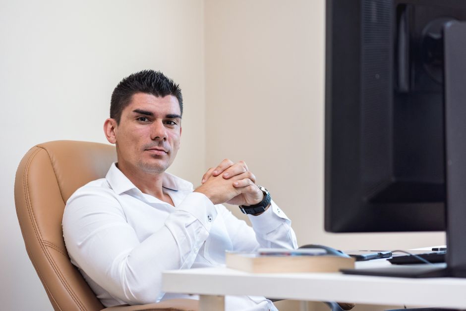 Businessman in white shirt sitting confidently at his desk with hands clasped, next to a computer monitor.
