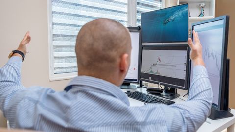 A stock trader in an office raises his hands in celebration while monitoring multiple screens with financial charts.