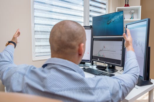 A stock trader in an office raises his hands in celebration while monitoring multiple screens with financial charts.