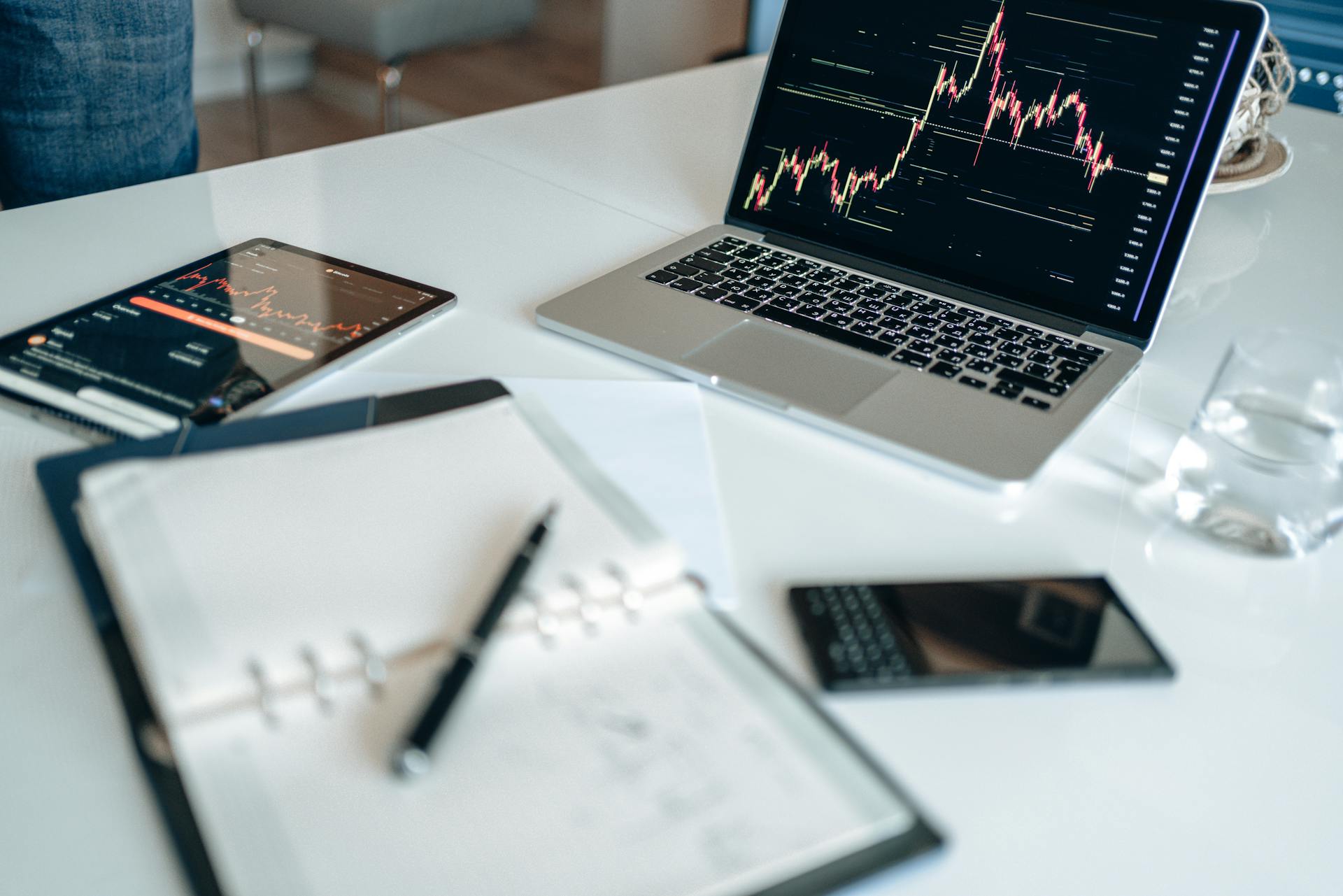 Modern office desk with laptop displaying financial graphs, tablet, and open notebook.