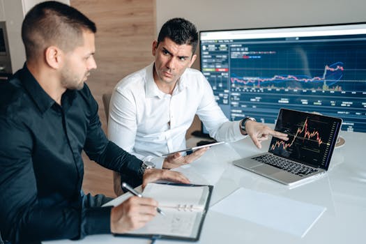 Two businessmen discuss financial charts on laptops in an office setting.
