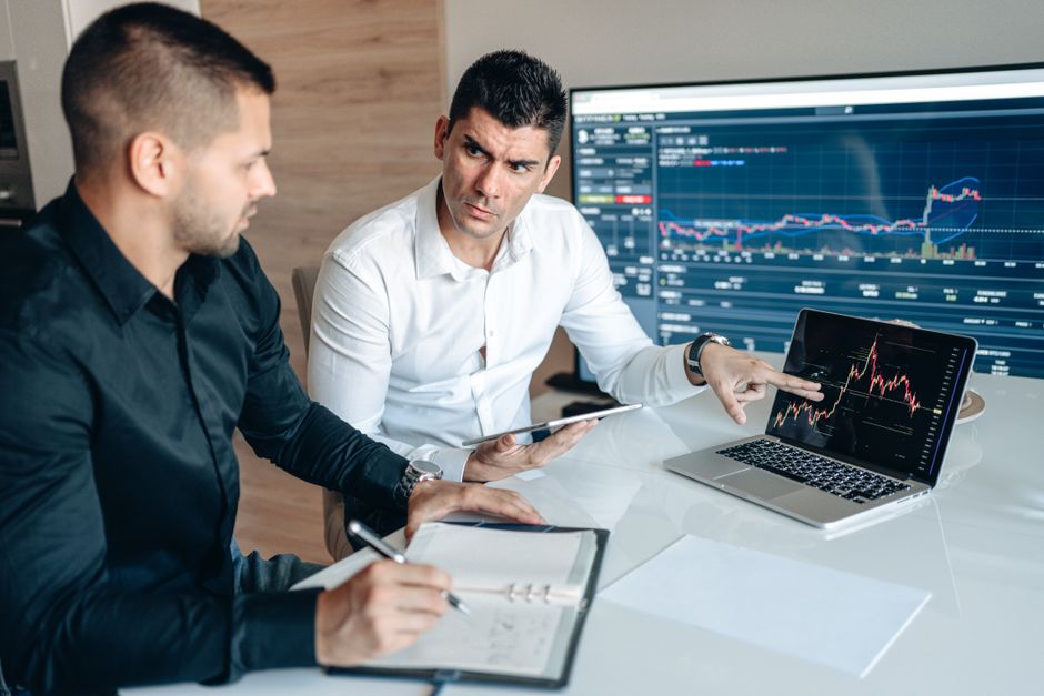Two businessmen discuss financial charts on laptops in an office setting. Two businessmen discuss financial charts on laptops in an office setting.