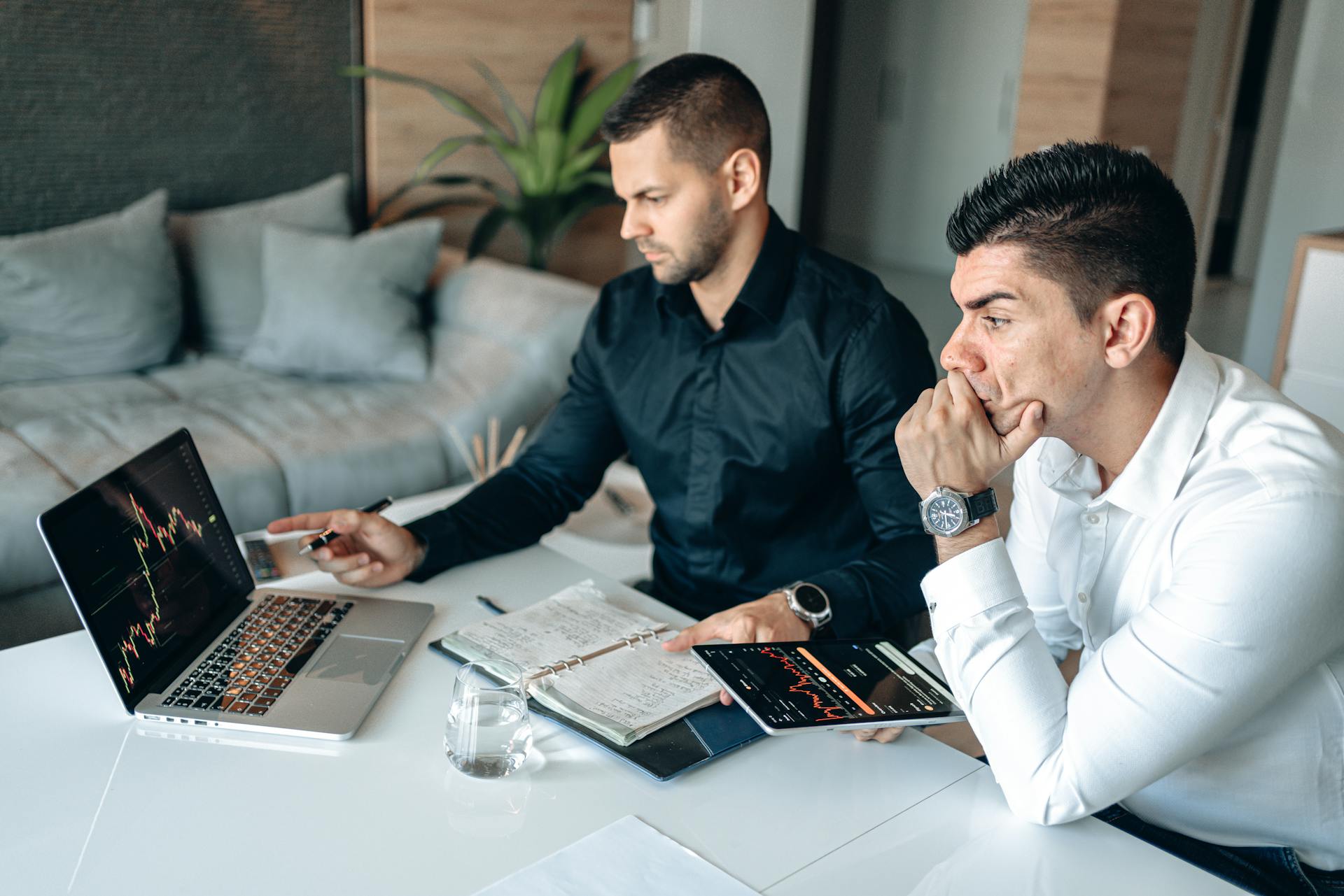 Two men studying financial graphs on a laptop and digital tablet in a modern office setting.