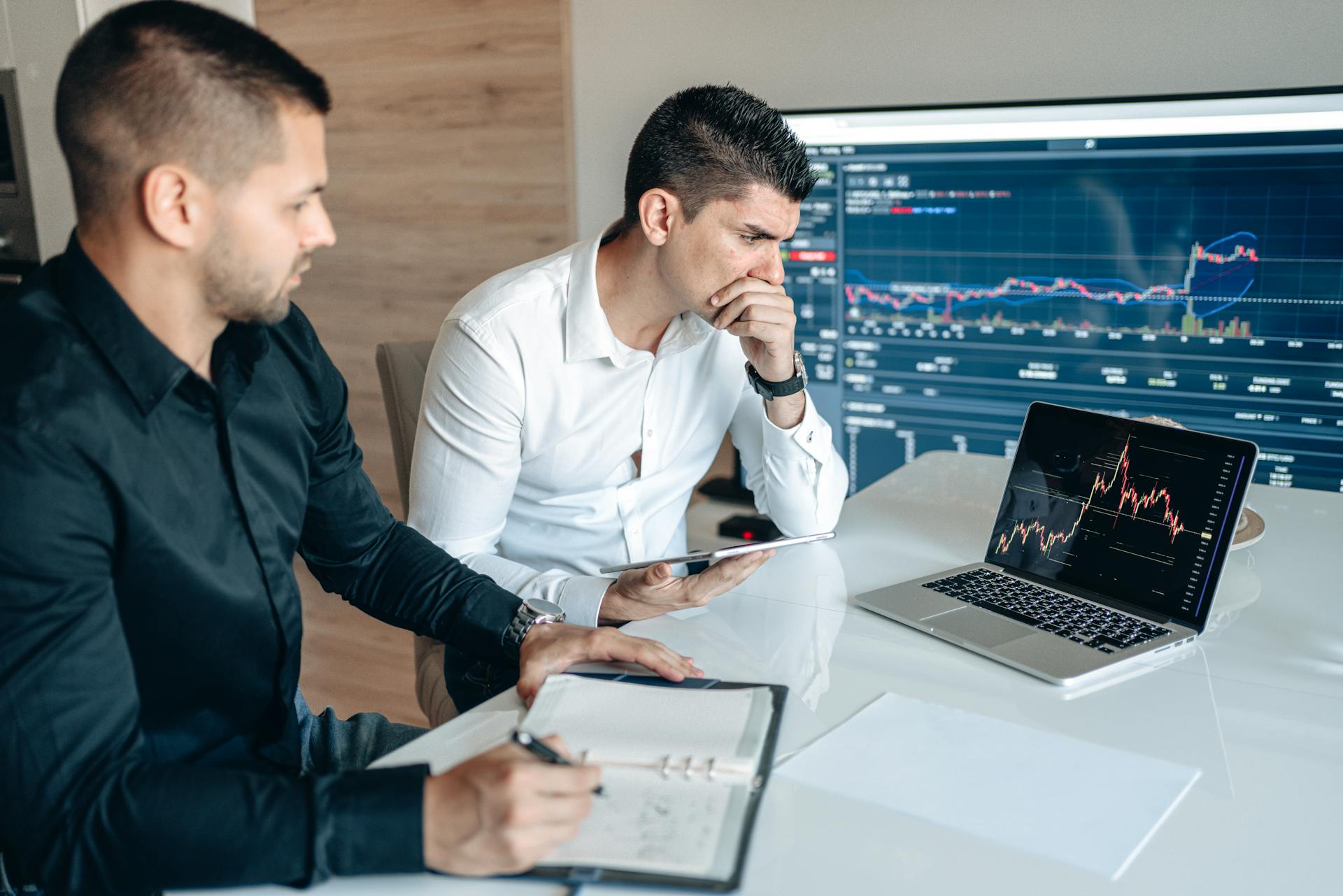 Two businessmen analyzing stock market data on laptops and tablets in an office setting.