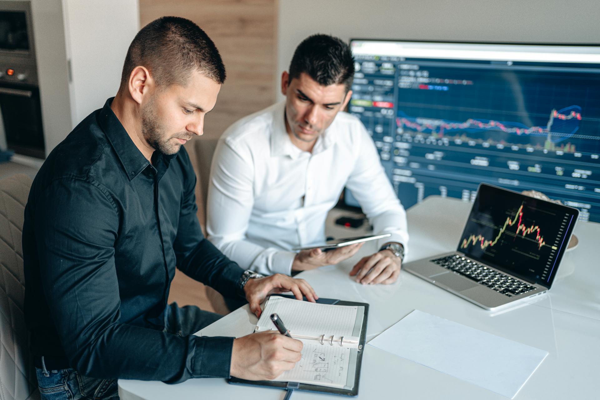 Two businessmen analyzing financial data with digital devices and charts in an office setting.