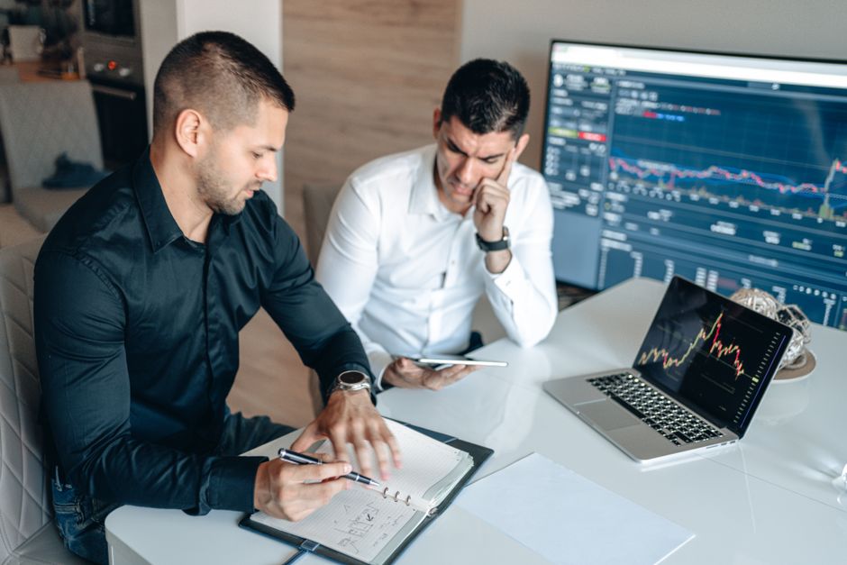 Two professionals discussing market trends with digital devices and charts in an office setting. Two professionals discussing market trends with digital devices and charts in an office setting.