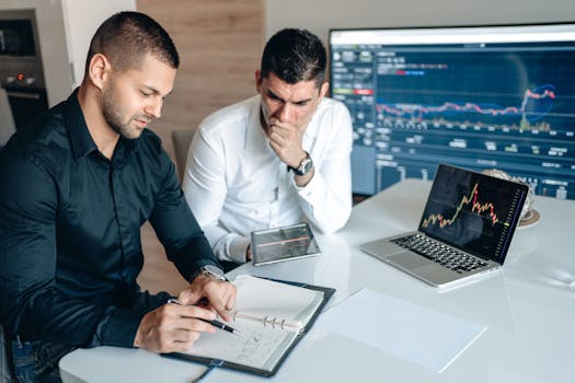 Two businessmen reviewing financial charts on a laptop and tablet in a modern office setting.