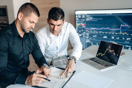 Two businessmen analyze financial charts on screens and notebook in office setting.