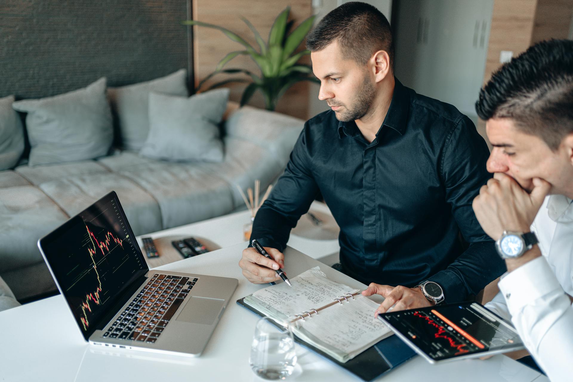 Two businessmen analyzing financial charts on laptop and tablet in office setting.