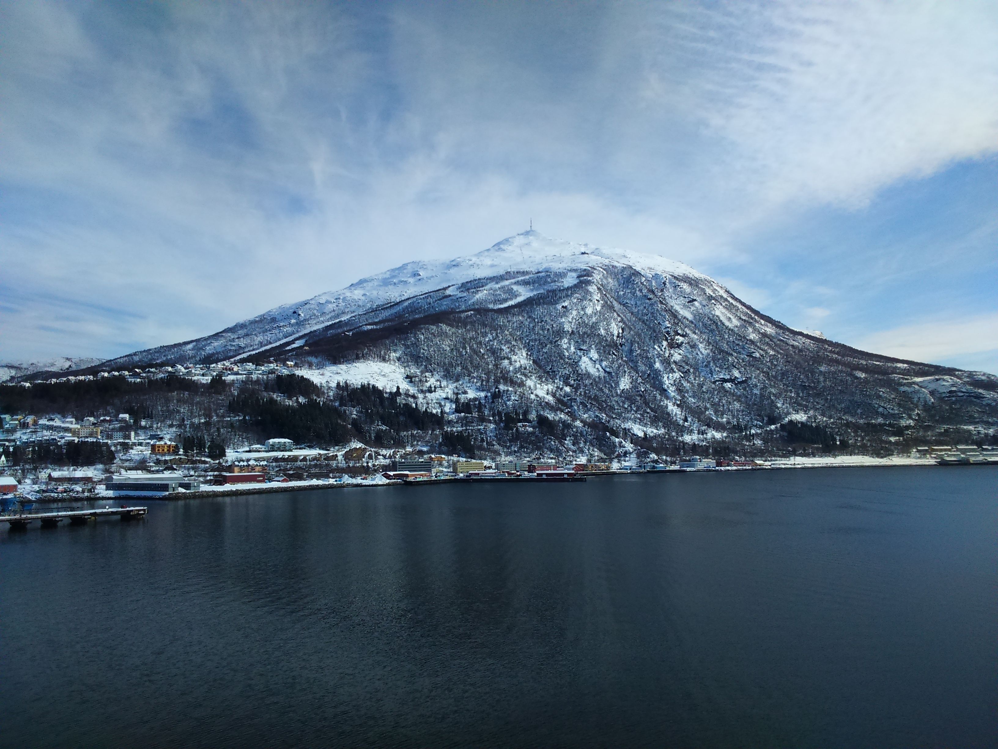 A serene winter scene of a snow-capped mountain reflected in a calm fjord under cloudy skies.