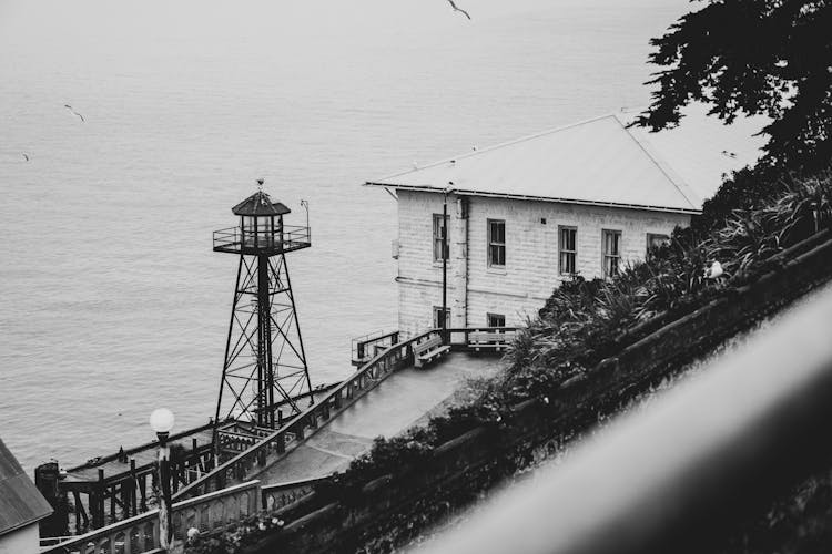 Grayscale Photo Of A House And Guard Tower Beside The Sea