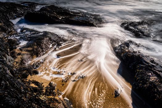 From above of foamy waves of powerful ocean washing sandy beach with rocky formations and seaweed on sunny day