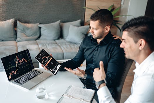 Two businessmen discuss digital trading insights with laptop and tablet in a modern office setting.