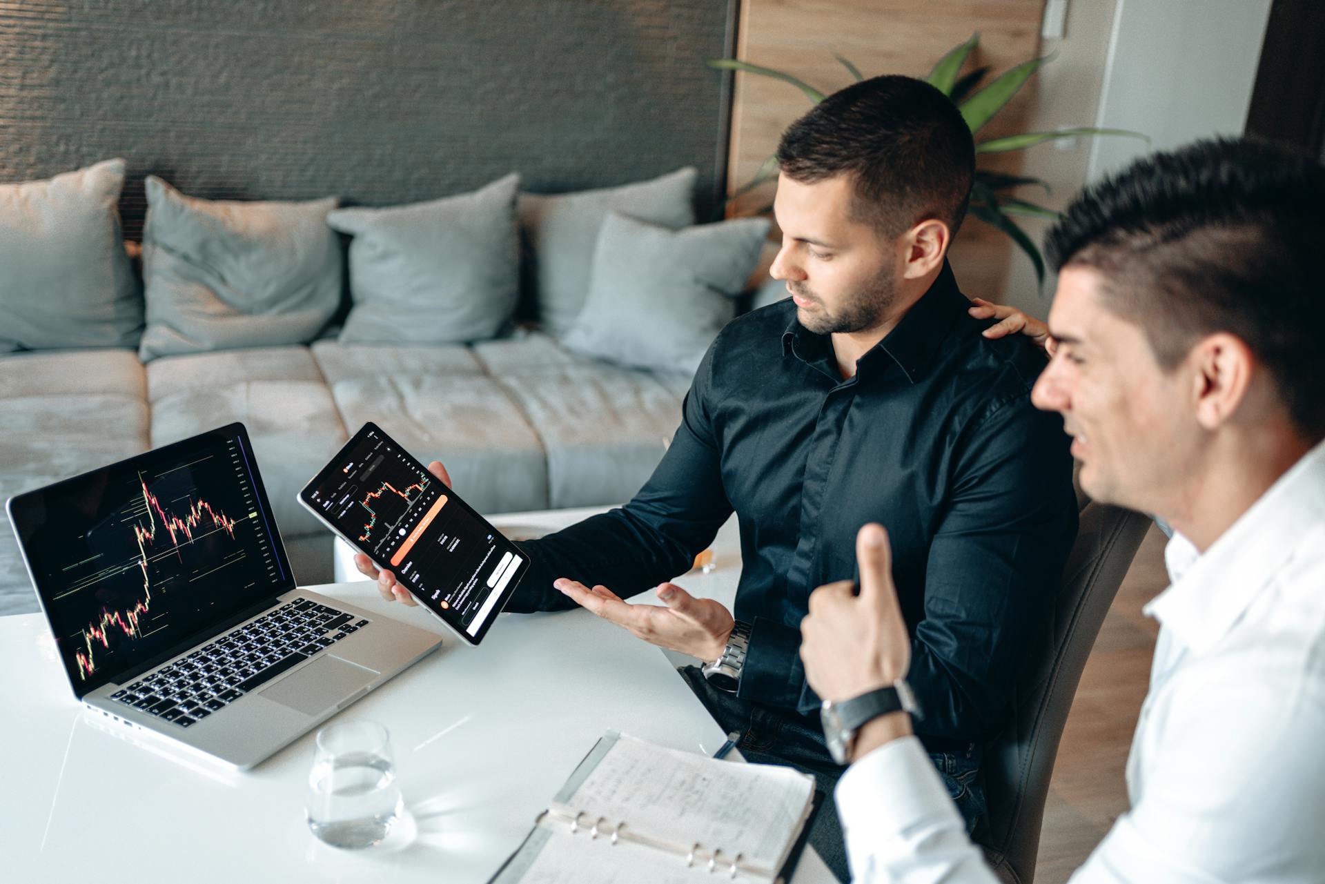 Two businessmen discuss digital trading insights with laptop and tablet in a modern office setting.