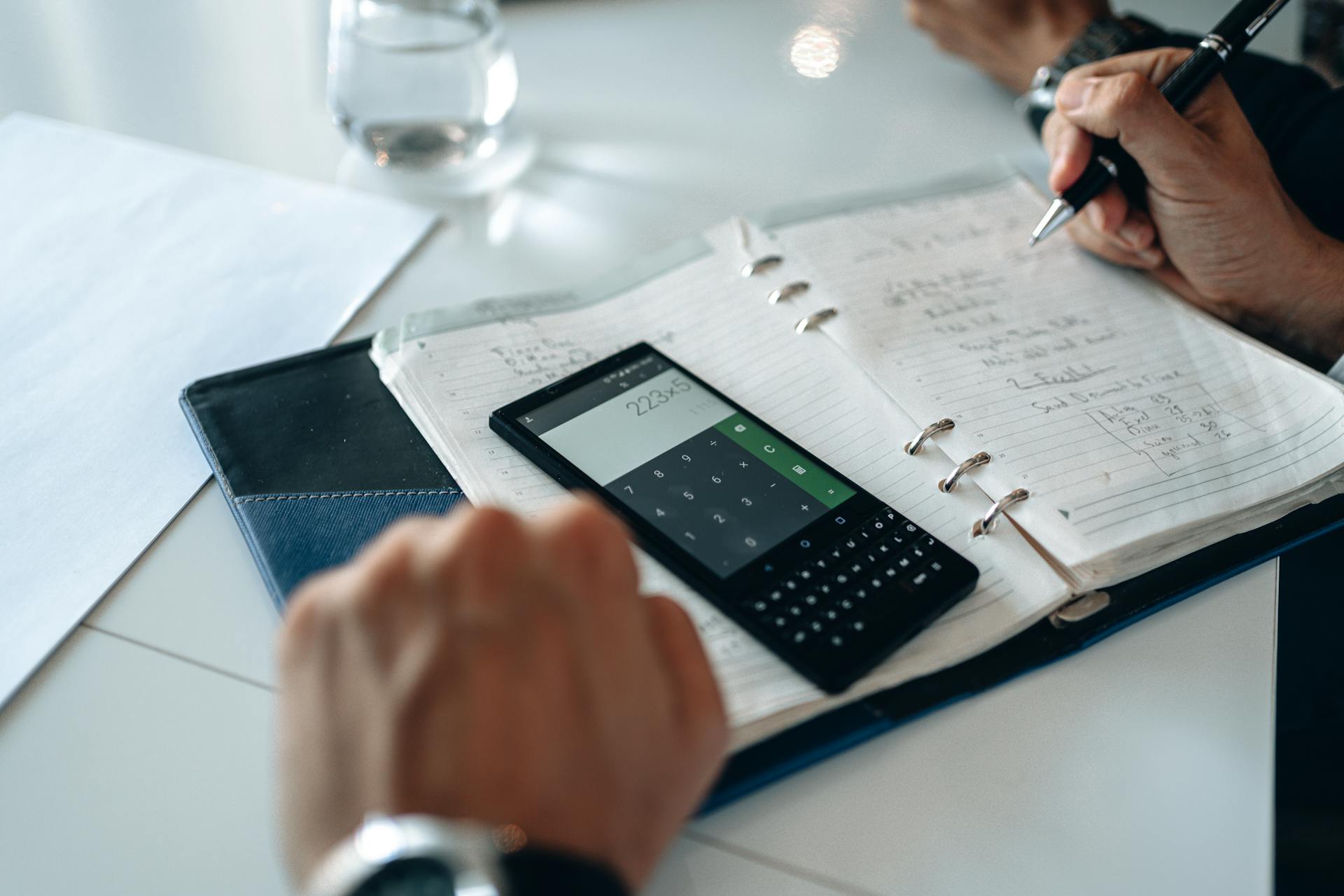 Close-up of hands writing in notebook with smartphone calculator on the table.