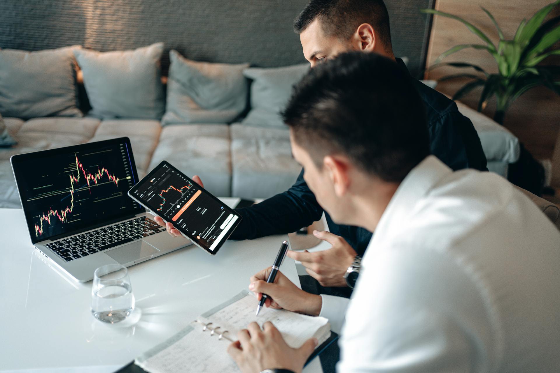 Two men discussing market trends using a tablet and laptop in a modern office.