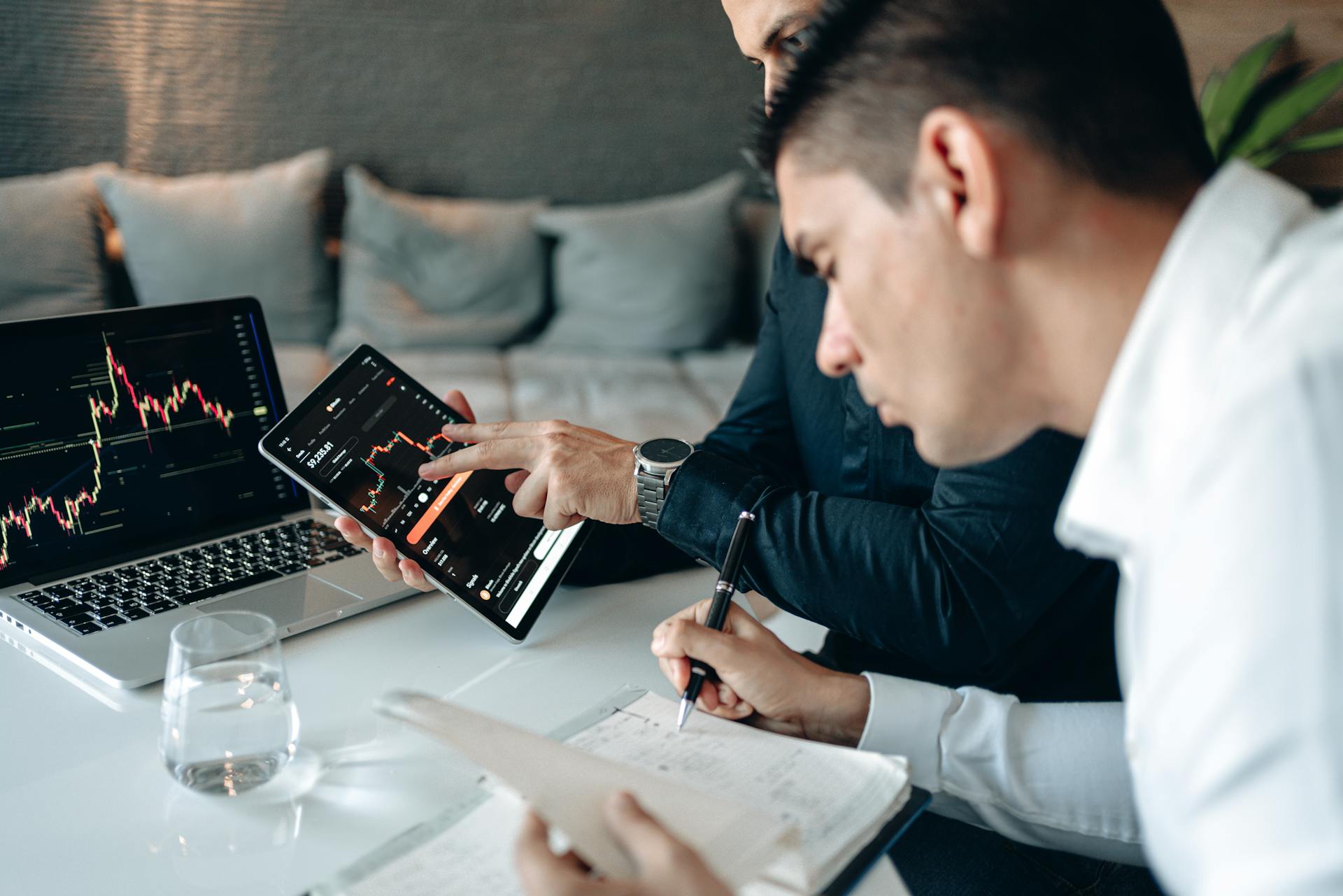 Two businessmen in a meeting analyzing financial data with digital devices and taking notes.