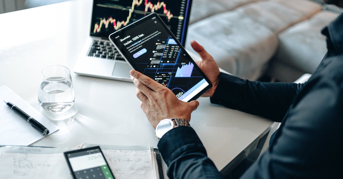 Person holding a tablet showing market data, with laptop and pen on white table.