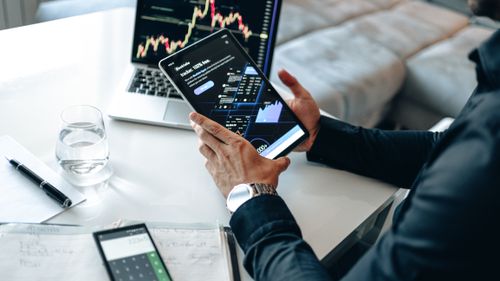 Free Person holding a tablet showing market data, with laptop and pen on white table. Stock Photo