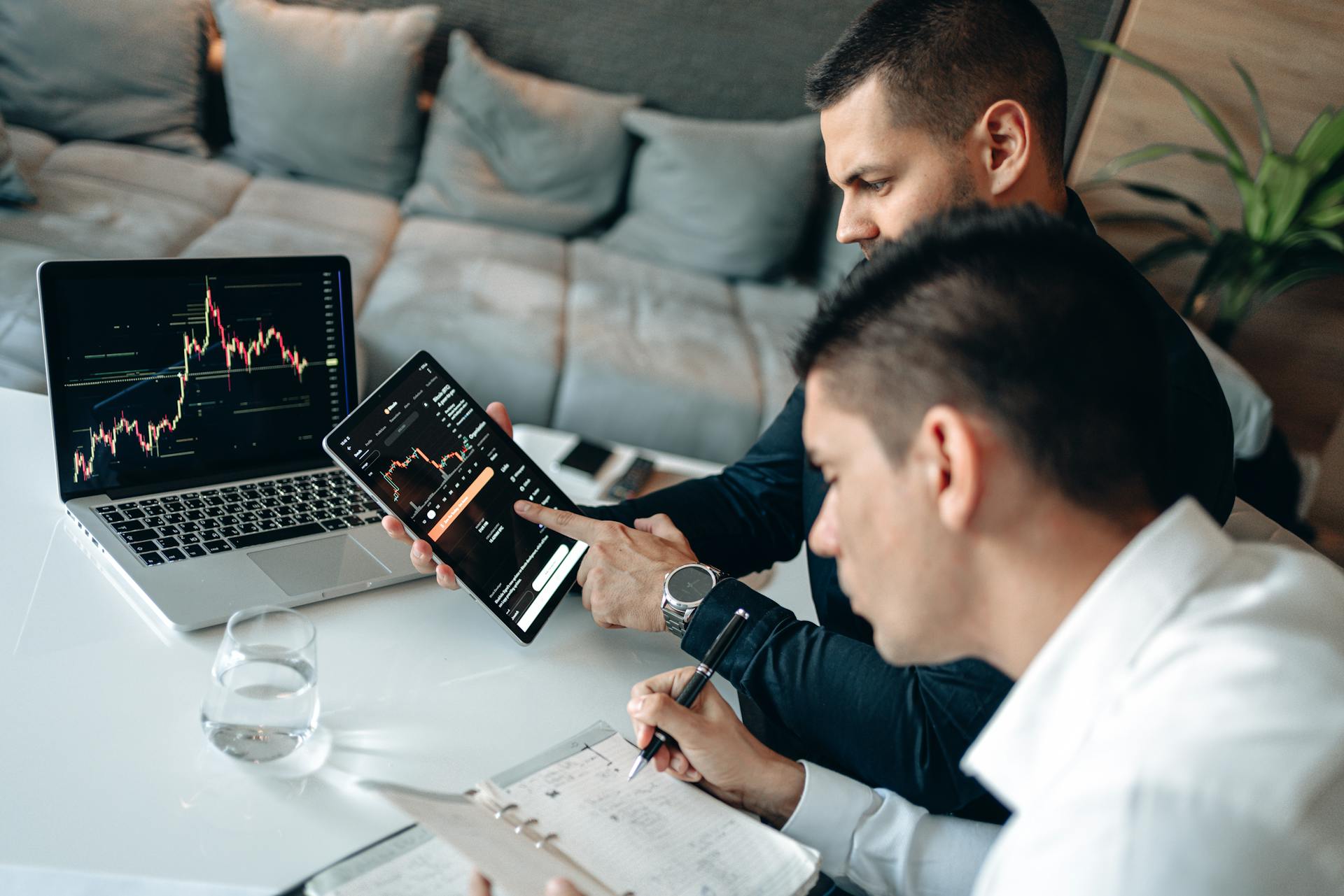 Two businessmen discussing financial charts on devices at a modern indoor setting.