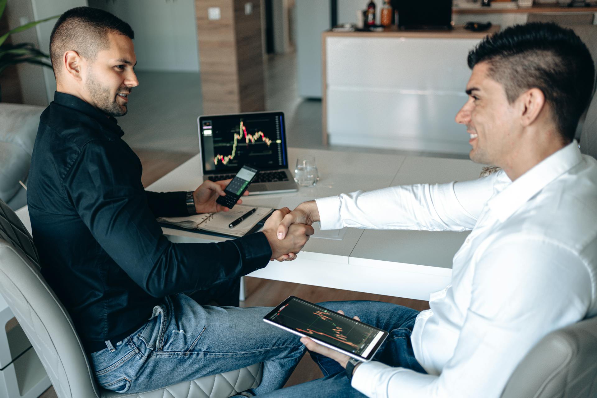 Two men shaking hands over a successful business deal with trading charts displayed.