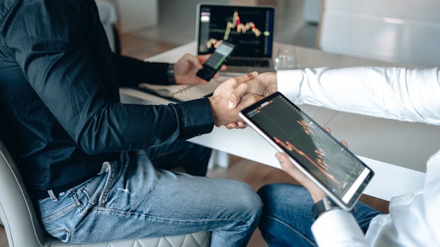 Two business professionals shaking hands with financial graphs on digital devices.