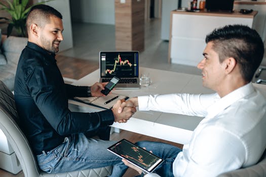 Two businessmen shake hands over a successful stock trading agreement using technology in a modern office.