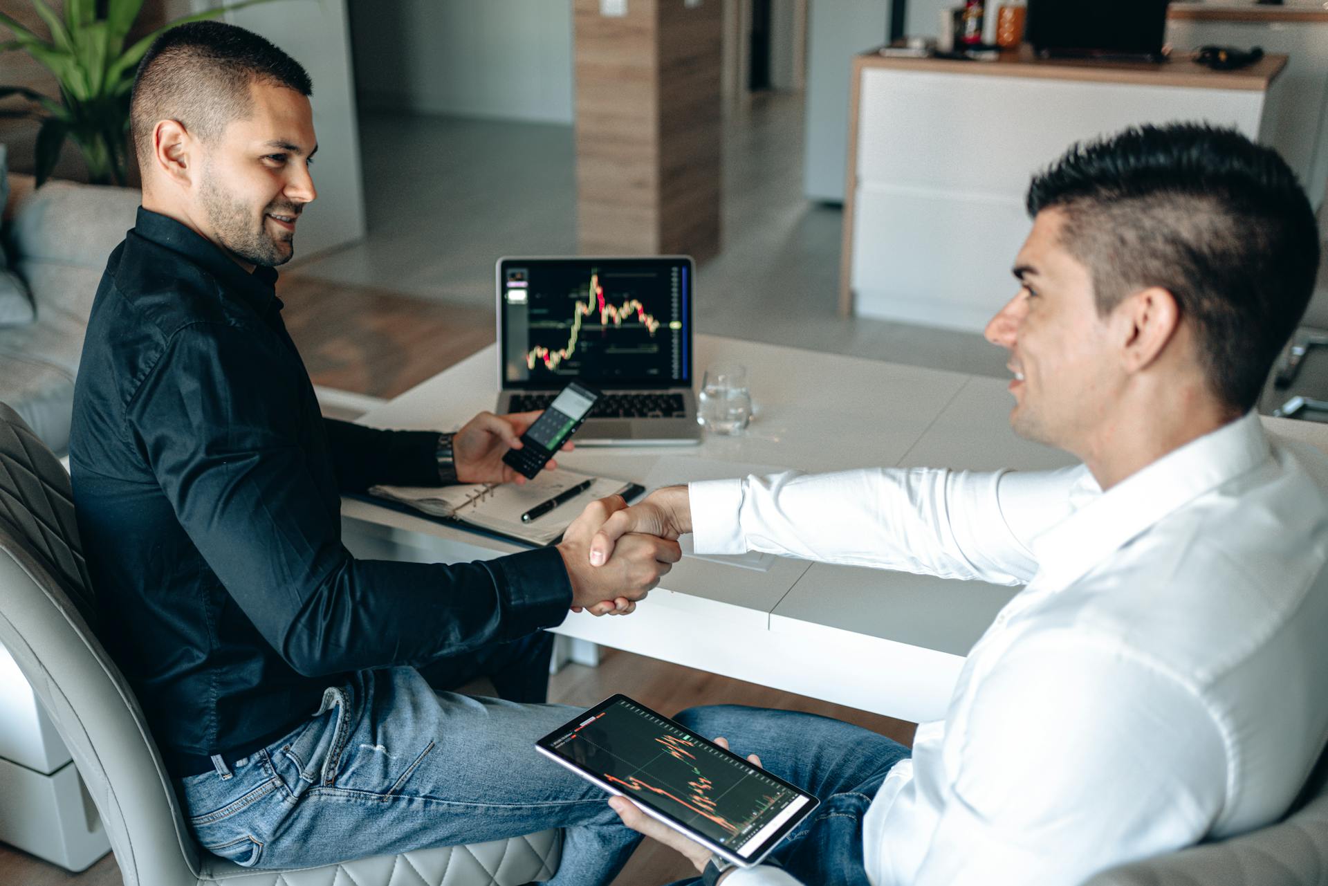 Two businessmen shake hands over a successful stock trading agreement using technology in a modern office.