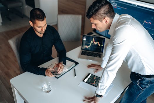 Two businessmen analyzing cryptocurrency charts on gadgets in an office setting.