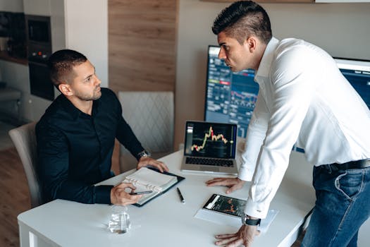 Two businessmen discuss stock market data on screens in a modern office.