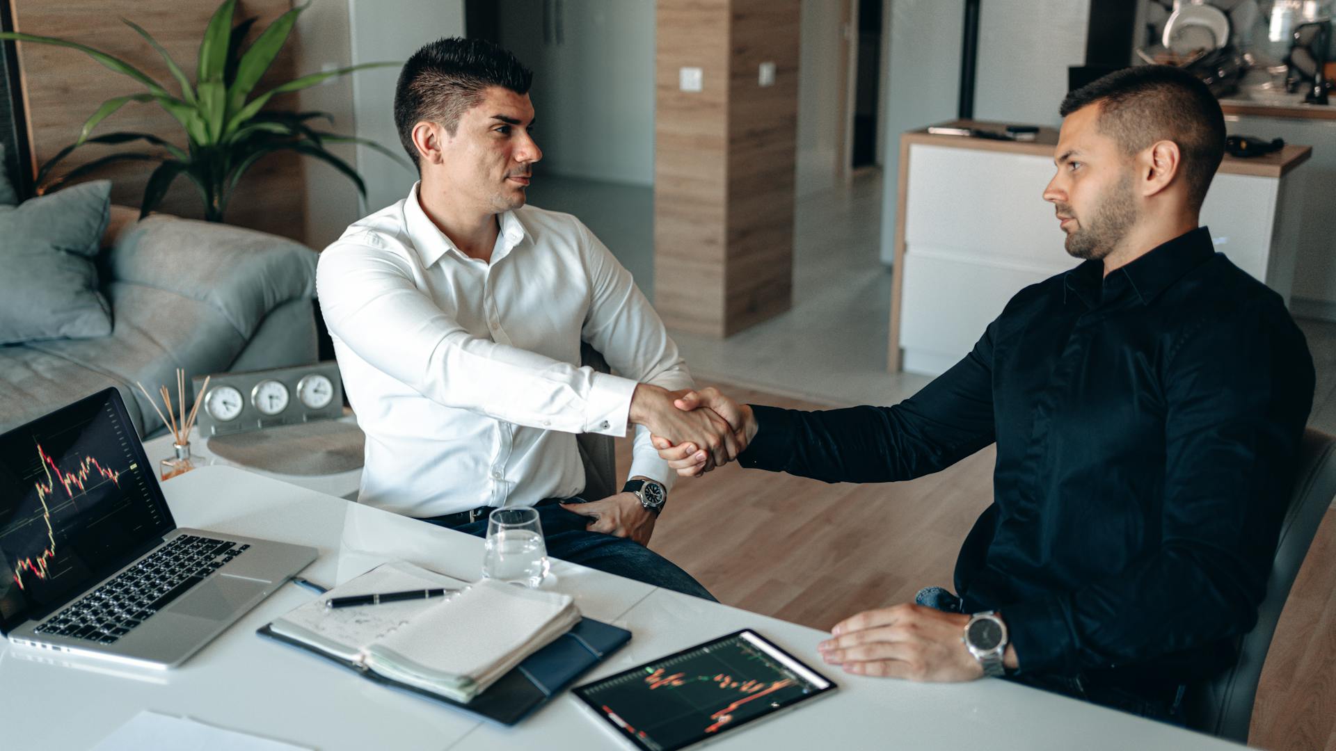 Two businessmen shake hands over financial charts and gadgets, symbolizing a successful deal.