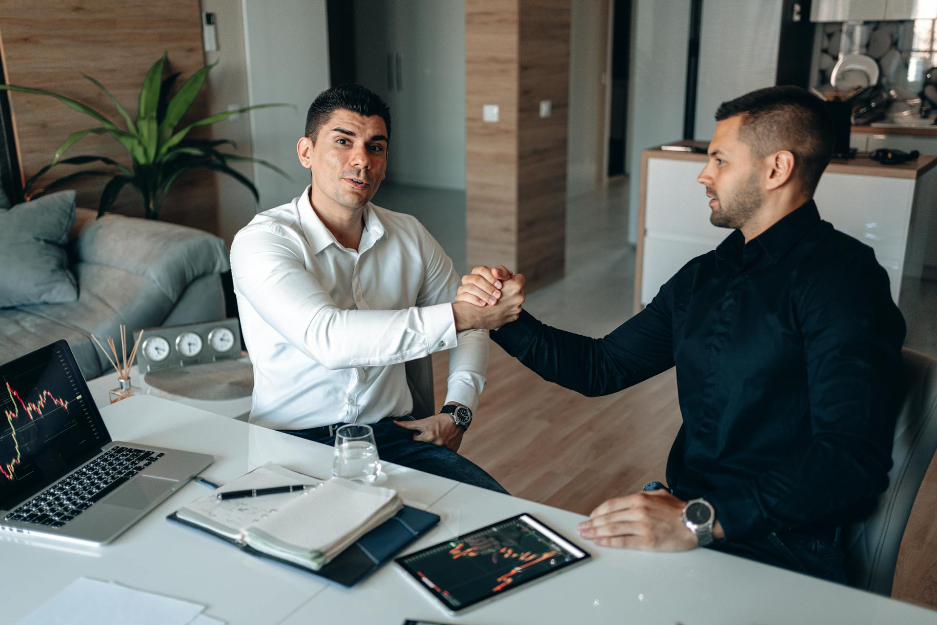 Two businessmen shake hands during a meeting in a modern office setting with digital equipment.