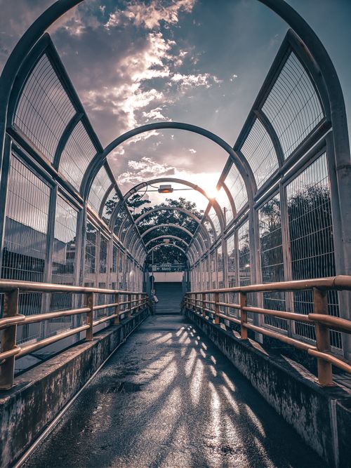 Free A covered pedestrian bridge with metal railings under a cloudy sky, featuring soft light and shadows. Stock Photo