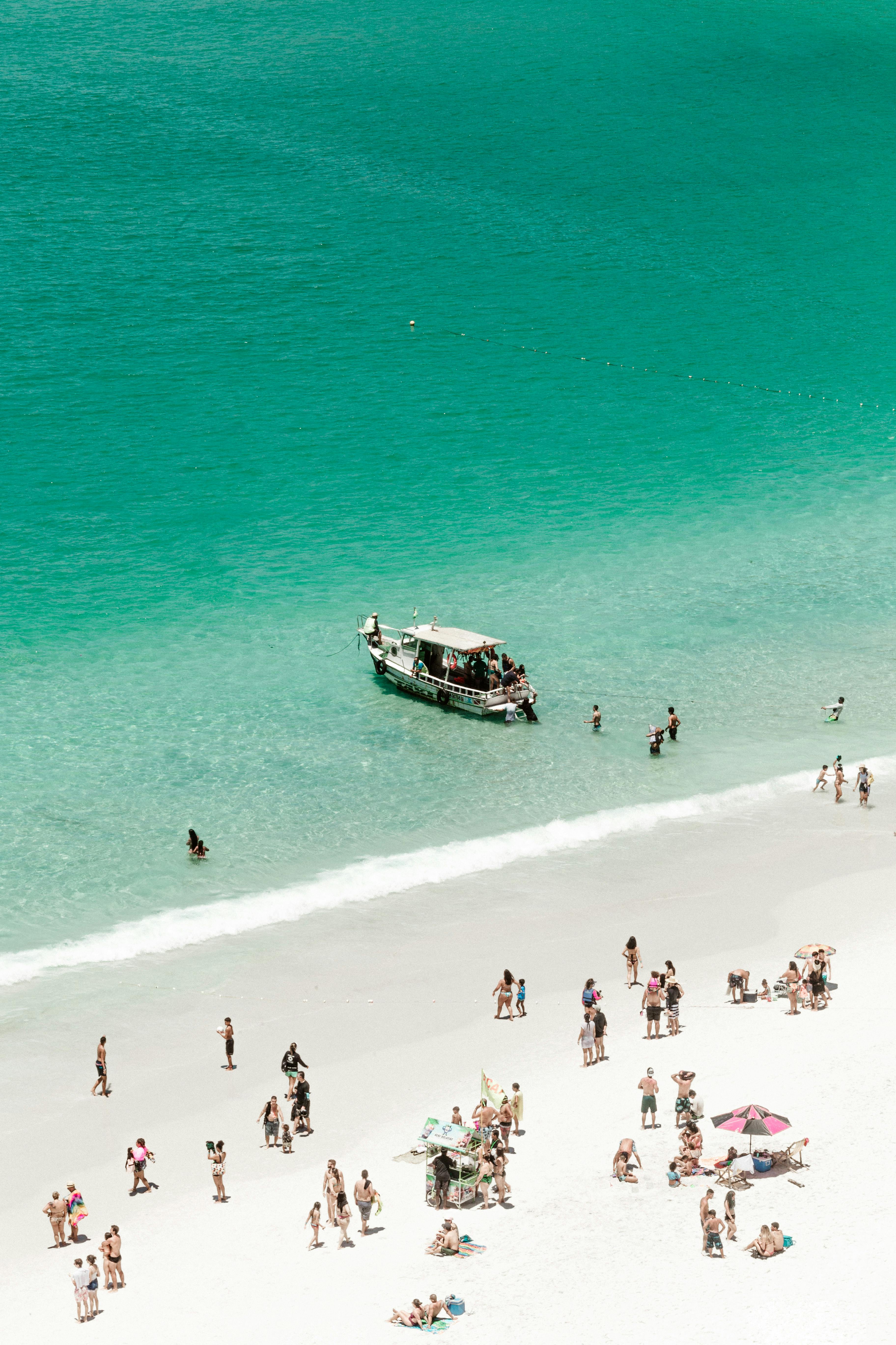 From above of anonymous travelers relaxing on sandy beach near waving turquoise ocean with floating yacht