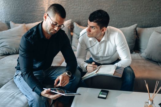 Two men in a home office discussing business topics using a tablet and notebook.